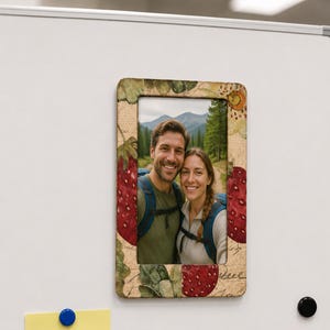 Puede incluir: Un marco de fotos rectangular con un fondo beige texturizado y un patrón impreso de fresas rojas y hojas verdes. El marco contiene una fotografía de dos personas con mochilas en un entorno de bosque montañoso.