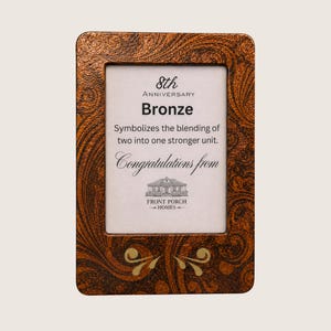 Puede incluir: Un marco de fotos rectangular con un acabado de bronce texturizado y patrones oscuros en espiral. El texto interior dice: 8th Anniversary Bronze, Symbolizes the blending of two into one stronger unit. Congratulations from Front Porch Homes.