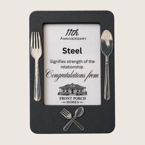 Puede incluir: Un marco rectangular negro con un inserto blanco que contiene el texto 11th Anniversary, Steel, Signifies strength of the relationship, Congratulations from y Front Porch Homes. Un tenedor y una cuchara plateados están fijados a los lados.