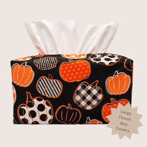 May include: A rectangular tissue box cover featuring a black background with a repeating pattern of orange and white pumpkins in various styles, including polka dots, stripes, and buffalo check. White tissues extend from the top opening.