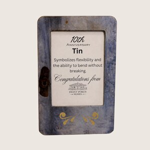 Puede incluir: Un marco rectangular de metal azul y gris jaspeado con un pestillo de metal oscuro en el lado izquierdo. El inserto central dice: 10th Anniversary Tin. Symbolizes flexibility and the ability to bend without breaking. Congratulations from Front Porch Homes.