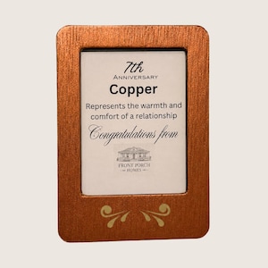Puede incluir: Un marco rectangular de color cobre con un acabado metálico cepillado y detalles dorados en forma de volutas en la parte inferior. El inserto dice: 7th Anniversary Copper, Represents the warmth and comfort of a relationship, Congratulations from Front Porch Homes.