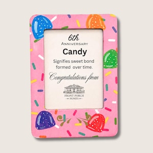 Puede incluir: Un marco de fotos rectangular de color rosa decorado con caramelos pintados y chispas de colores. El inserto presenta el texto: 6th Anniversary Candy Signifies sweet bond formed over time. Congratulations from Front Porch Homes.