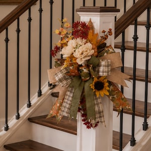 May include: A decorative autumn floral arrangement featuring cream peonies, dark red chrysanthemums, and a yellow sunflower. The display is accented with brown berries, pinecones, and a multi-layered bow made of hessian, plaid, and leaf-patterned ribbons.