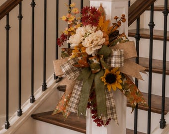 Op de afbeelding: Een decoratief herfstbloemstuk met crèmekleurige pioenen, donkerrode chrysanten en een gele zonnebloem. Het arrangement is versierd met bruine bessen, dennenappels en een meerlaagse strik van jute, ruitjesstof en linten met een bladmotief.