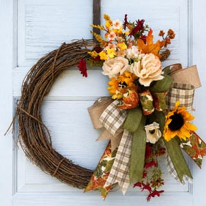 May include: A circular wreath made of brown twisted twigs decorated with artificial flowers in shades of yellow, cream, and deep red, accented by orange maple leaves, green foliage, and a multi-layered bow featuring plaid and solid olive green ribbons.
