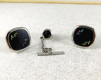 May include: A vintage set of cufflinks and tie tack. The set features square cufflinks and a round tie tack with a black background and colorful accents. The metal has a silver finish.