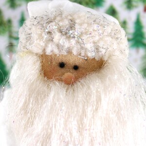 May include: Close-up of a Santa Claus doll with a white beard and hat. The doll's face is made of tan fabric with black eyes and a small pink nose. The hat and beard have a fluffy texture with silver glitter accents. Christmas decor.