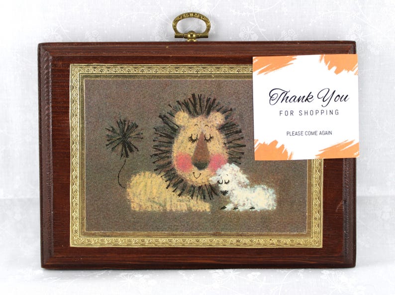 May include: A framed artwork featuring a lion and a lamb. The lion has a brown mane and is lying down with its eyes closed. The lamb is white and is standing next to the lion. The artwork is on a brown background and has a gold frame.