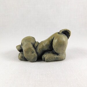 May include: A small, grey, ceramic sculpture of a dog lying on its belly with its head turned to the side. The dog has a short tail and its ears are floppy.