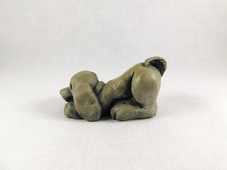 May include: A small, grey, ceramic sculpture of a dog lying on its belly with its head turned to the side. The dog has a short tail and its ears are floppy.