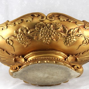 May include: An oval-shaped gold-colored bowl featuring intricate relief carvings of grape clusters and vine leaves along the exterior. The bowl rests on a pedestal base with decorative scrollwork and has a light-colored felt underside.