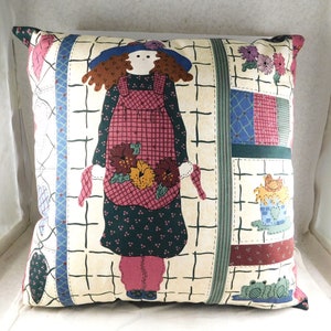 May include: A square throw pillow with a patchwork design featuring a girl in a red and white gingham dress holding flowers. The background is a cream color with a grid pattern.