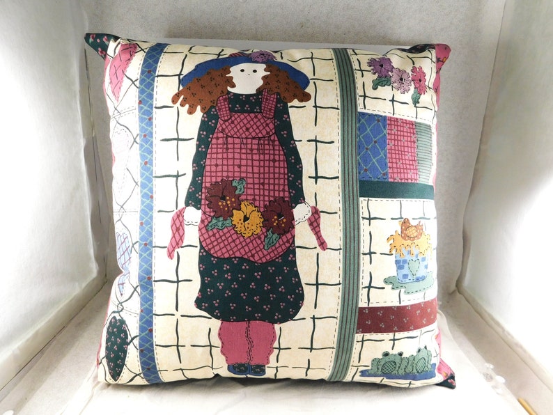 May include: A square throw pillow with a patchwork design featuring a girl in a red and white gingham dress holding flowers. The background is a cream color with a grid pattern.