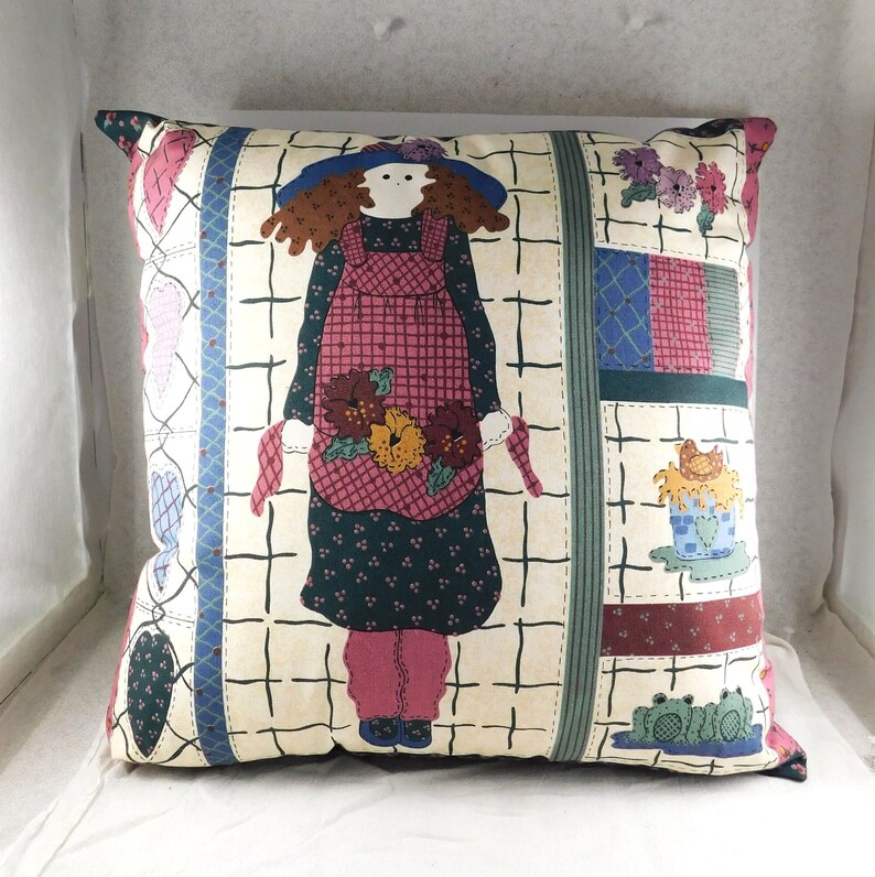 May include: A square throw pillow with a cream background and a patchwork design. The center features a cartoon girl with red hair wearing a blue hat and a pink and red dress. The girl is holding a bouquet of flowers. The pillow has a green, blue, and red border.