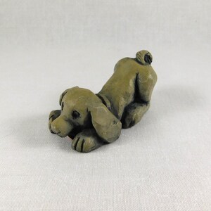 May include: A small, hand-carved, green ceramic figurine of a dog lying down with its head turned to the side. The dog has a short tail and is sculpted in a playful pose.