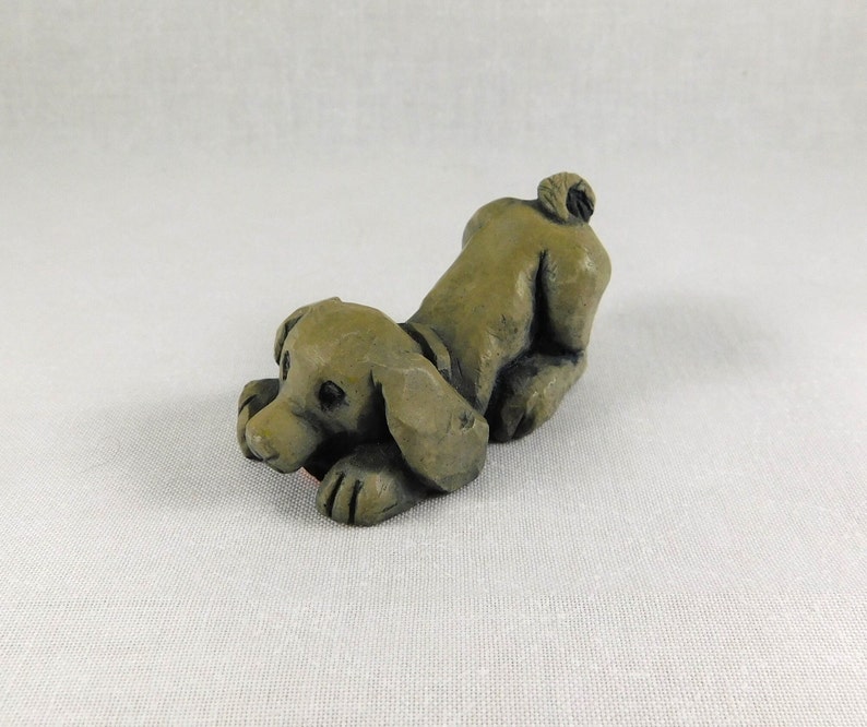 May include: A small, hand-carved, green ceramic figurine of a dog lying down with its head turned to the side. The dog has a short tail and is sculpted in a playful pose.