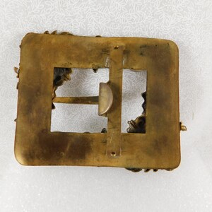 K&ouml;nnte beinhalten: Eine goldfarbene G&uuml;rtelschnalle mit einem verzierten Design. Die Schnalle hat eine rechteckige Form mit einer kleineren rechteckigen &Ouml;ffnung in der Mitte. Die Schnalle ist mit komplizierten Blumenmustern verziert.