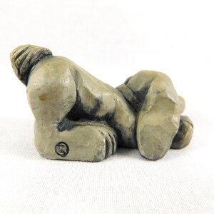 May include: A small, grey ceramic sculpture of a dog lying on its side with its tail curled up. The sculpture is detailed with textured fur and a slightly distressed finish.