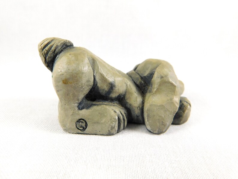 May include: A small, grey ceramic sculpture of a dog lying on its side with its tail curled up. The sculpture is detailed with textured fur and a slightly distressed finish.