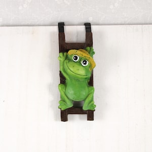 May include: A green frog figurine wearing a yellow straw hat, climbing a brown ladder. The frog has large, round eyes and a smiling expression. The ladder is attached to a white surface, creating a whimsical garden decoration.