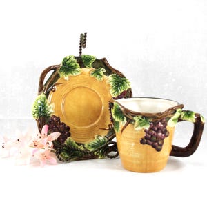 May include: A ceramic pitcher and saucer set with a grape and leaf design. The pitcher and saucer are yellow with brown accents and feature green leaves and purple grapes. The handle of the pitcher is brown.