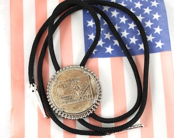 May include: A vintage bolo tie with a circular silver-toned pendant featuring the text "NATIONAL SCOUT JAMBOREE 1977". The pendant is attached to a black braided cord with silver-toned tips. The background is an American flag.
