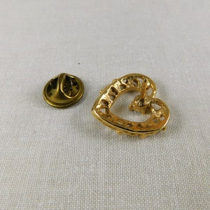 May include: Gold heart-shaped pin with a decorative design. The pin has a back closure with a safety pin.
