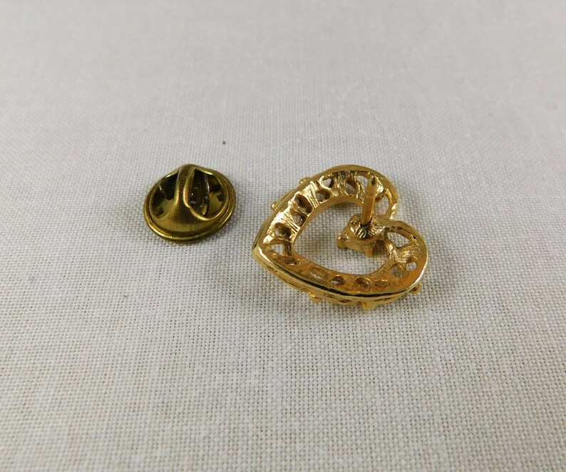 May include: Gold heart-shaped pin with a decorative design. The pin has a back closure with a safety pin.