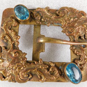 K&ouml;nnte beinhalten: Eine antike G&uuml;rtelschnalle aus Messing mit verzierten Blumenmustern und zwei blauen Glassteinen.