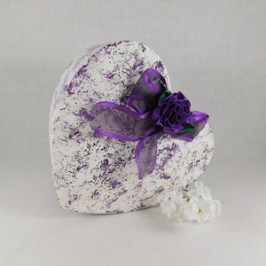 Op de afbeelding: Een witte hartvormige doos met een marmerpatroon in paars en wit. De doos is versierd met een paars lint en een paarse bloem.