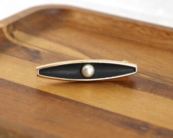 May include: A gold-toned metal brooch featuring an elongated, pointed oval shape with a matte black inlay and a single centered white pearl. The brooch has a smooth, polished metallic edge that frames the dark interior and the round pearl.