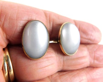 May include: A pair of oval-shaped earrings with a pearly, light gray center stone set in a gold-colored metal frame. The earrings are displayed against a neutral background.