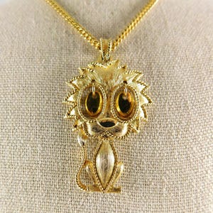 May include: A gold-tone necklace with a lion pendant. The lion has large, round, amber-colored eyes and a detailed mane.