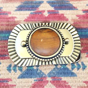 May include: Rectangular brass belt buckle with a central, round, brown stone. The buckle has a decorative, ridged border and two small, round rivets. The background is a colorful, patterned textile.