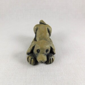 May include: A small, hand-carved wooden sculpture of a brown dog lying down with its head resting on its paws. The dog has a simple, stylized design with large eyes and a short tail.