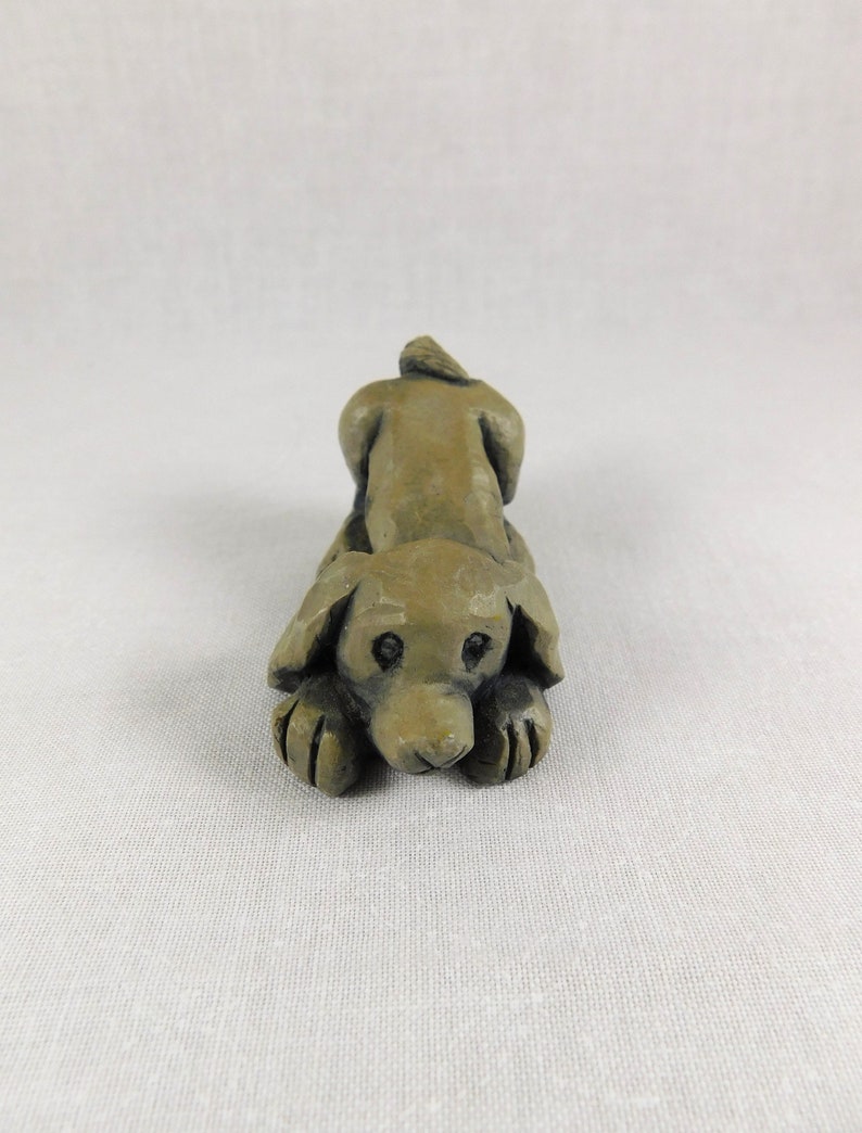 May include: A small, hand-carved wooden sculpture of a brown dog lying down with its head resting on its paws. The dog has a simple, stylized design with large eyes and a short tail.