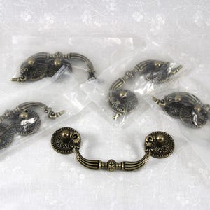 May include: A set of five antique brass-toned metal drawer pulls, each featuring a curved handle attached to two circular backplates with intricate embossed detailing. Four of the pulls are individually wrapped in clear plastic packaging.