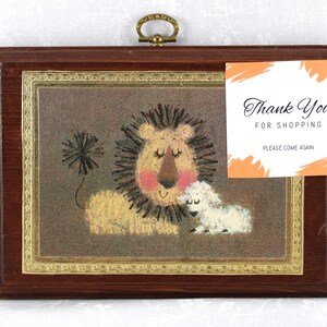 May include: A framed artwork with a lion and a lamb. The lion has a brown mane and is lying down with its eyes closed. The lamb is white and is standing next to the lion. The artwork is on a brown background and has a gold frame.