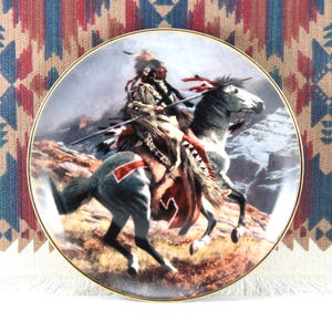 May include: Decorative plate featuring a Native American warrior on horseback, set against a mountainous backdrop. The plate has a gold rim and showcases detailed artwork with earthy tones and vibrant accents. The design evokes a sense of history and cultural heritage.
