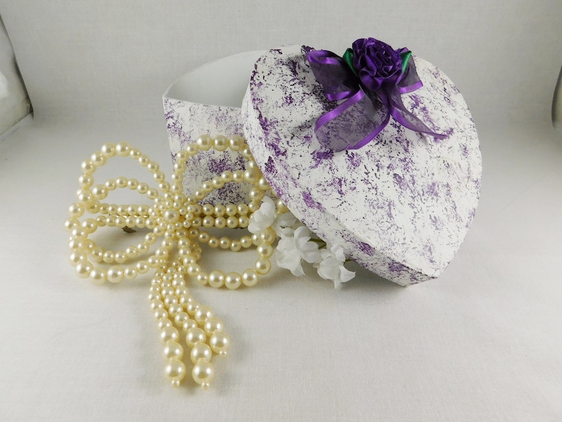 Op de afbeelding: Een witte hartvormige doos met een gespikkeld ontwerp in paars en wit. De doos is open en een parelketting in de vorm van een strik ligt ernaast. De doos is versierd met een paars lint en een paarse bloem.