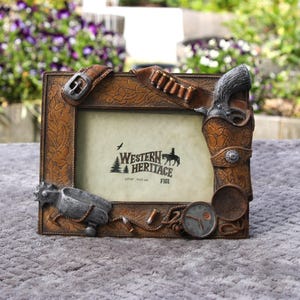 May include: A rectangular photo frame with a Western theme. The frame is brown with decorative elements including a gun holster, belt, and spurs. The frame has the text "Western Heritage" and is designed to hold a 4" x 6" photo.
