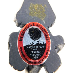 May include: A black volcanic ash sculpture of a bear with a red and white label that reads "Mt. St. Helen's Sculptures" and "Crafted by Hand with Genuine Volcanic Ash".