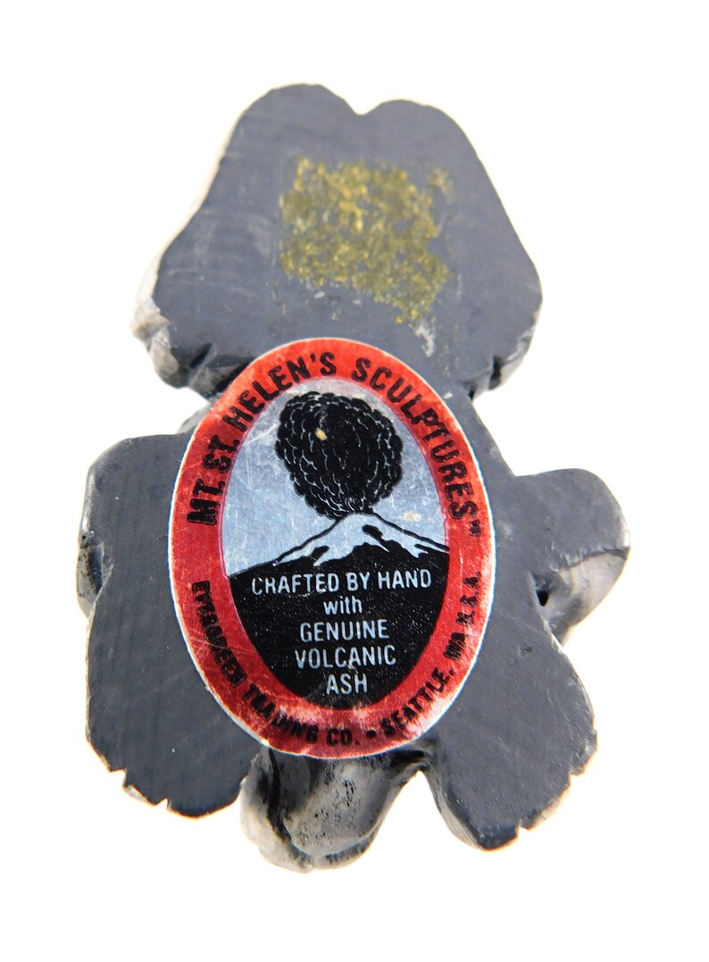 May include: A black volcanic ash sculpture of a bear with a red and white label that reads "Mt. St. Helen's Sculptures" and "Crafted by Hand with Genuine Volcanic Ash".