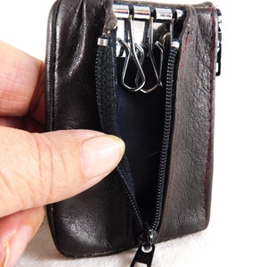 May include: A brown leather key holder with a zipper closure and a metal key ring with three hooks. The key holder is open and the zipper pull is visible.