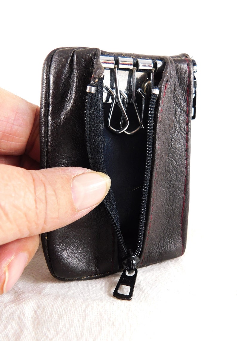 May include: A brown leather key holder with a zipper closure and a metal key ring with three hooks. The key holder is open and the zipper pull is visible.