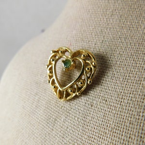 May include: A gold heart-shaped pendant with a green gemstone in the centre. The pendant is detailed with small beads around the edges.