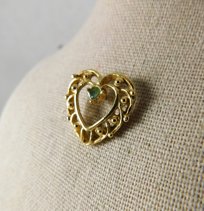 May include: A gold heart-shaped pendant with a green gemstone in the centre. The pendant is detailed with small beads around the edges.