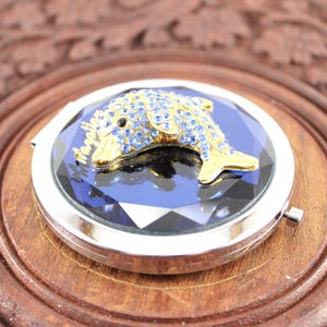 May include: A silver compact mirror with a blue crystal design and a gold dolphin with blue rhinestones on top.