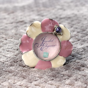 May include: A flower-shaped picture frame with a pink, cream, and silver design. The frame has a circular opening with the text "Jeweled Elegance" in a cursive font. A small purple gem is attached to the top.